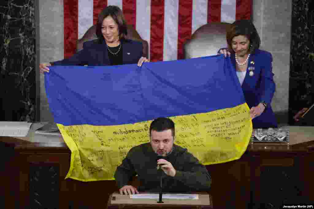 Vicepreședintele SUA, Kamala Harris (stânga) și președinta de atunci a Camerei Reprezentanților, Nancy Pelosi, reacționează în timp ce președintele ucrainean Volodimir Zelenski le prezintă legislatorilor americani un steag ucrainean cu semnăturile soldaților din trupele de pe frontul din Bahmut, în timp ce se adresează unei sesiuni comune a Congresului, la Washington, pe 21 decembrie 2022.