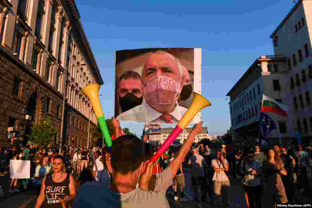 Një protestues mban në duar një fotografi të kryeministrit bullgar, i cili në këtë fotografi ka një maskë të vendosur të kartëmonedhës prej 500 eurove. Këta qytetarë morën pjesë në protestën anti-qeveritare në Sofja më 29 korrik, 2020, ku kërkuan dorëheqjen e qeverisë. (Nikolay Doychiov/AFP).